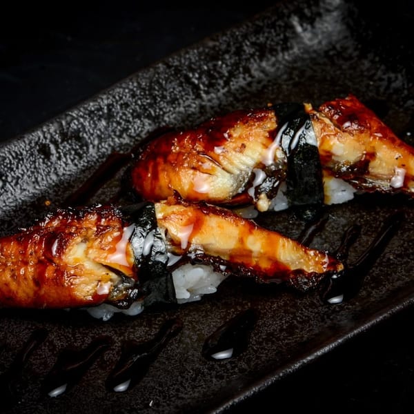 Two pieces of glistening unagi nigiri with sauce on a dark textured plate.