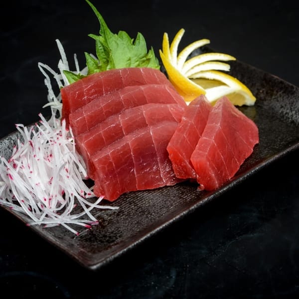 Sliced red tuna sashimi with shredded radish, green leaves, and lemon on a dark plate.