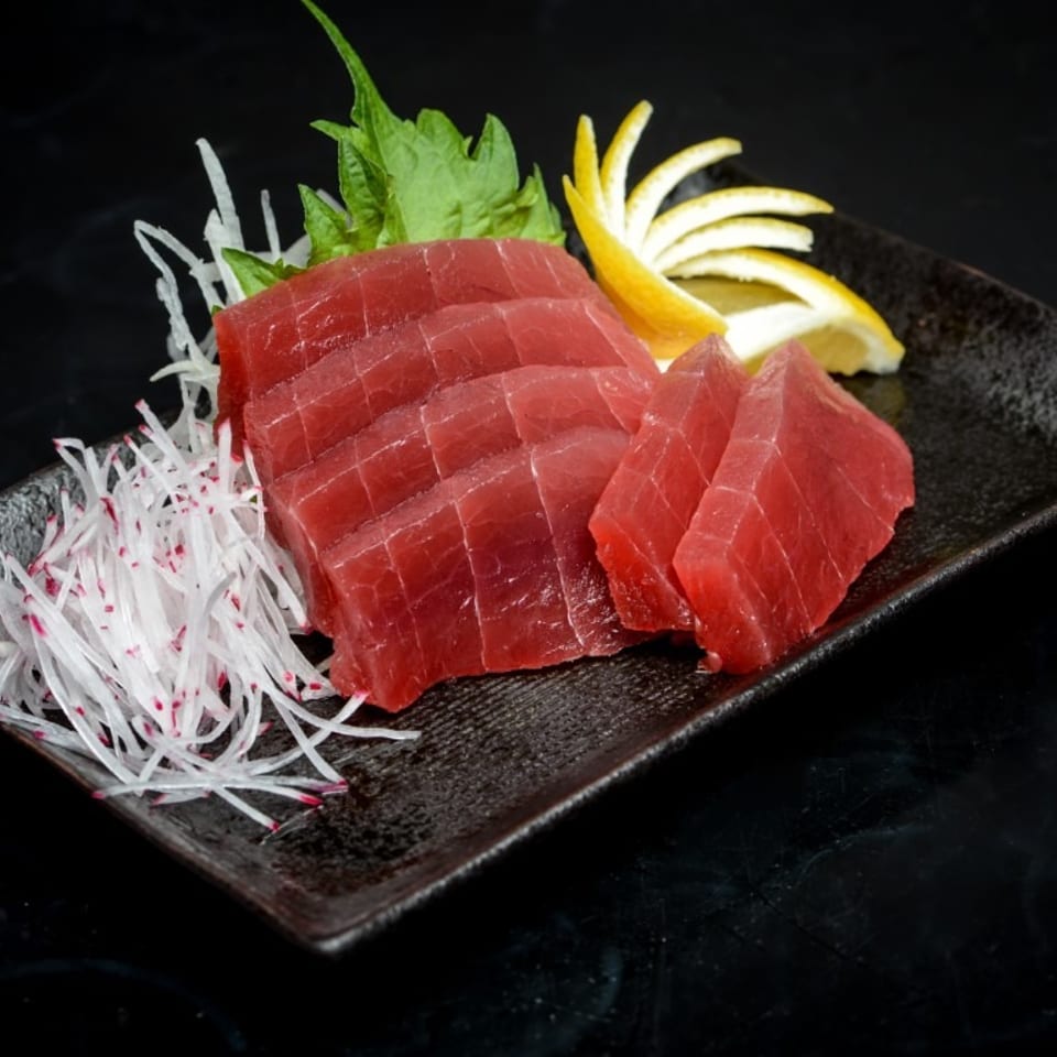 Sliced red tuna sashimi with shredded radish, green leaves, and lemon on a dark plate.